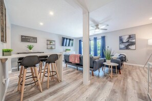 A modern living room with a sofa, a coffee table, and a TV. There is a dining area with a table and chairs. The room has wooden flooring and a ceiling fan. - Private Gulf Beach 3BR Townhome | Pools & Tennis (Destin)