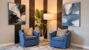Une pièce avec deux fauteuils bleus, un lampadaire, une plante en pot et des œuvres d’art abstrait accrochées aux murs.