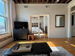 A room with a television, a bench, a small table with a remote, and a view through an open door to a bedroom with a bed and dresser.