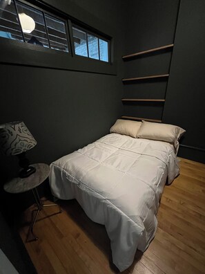 A small, dark-themed bedroom with a single bed, a nightstand, a lamp, and a window with blinds.