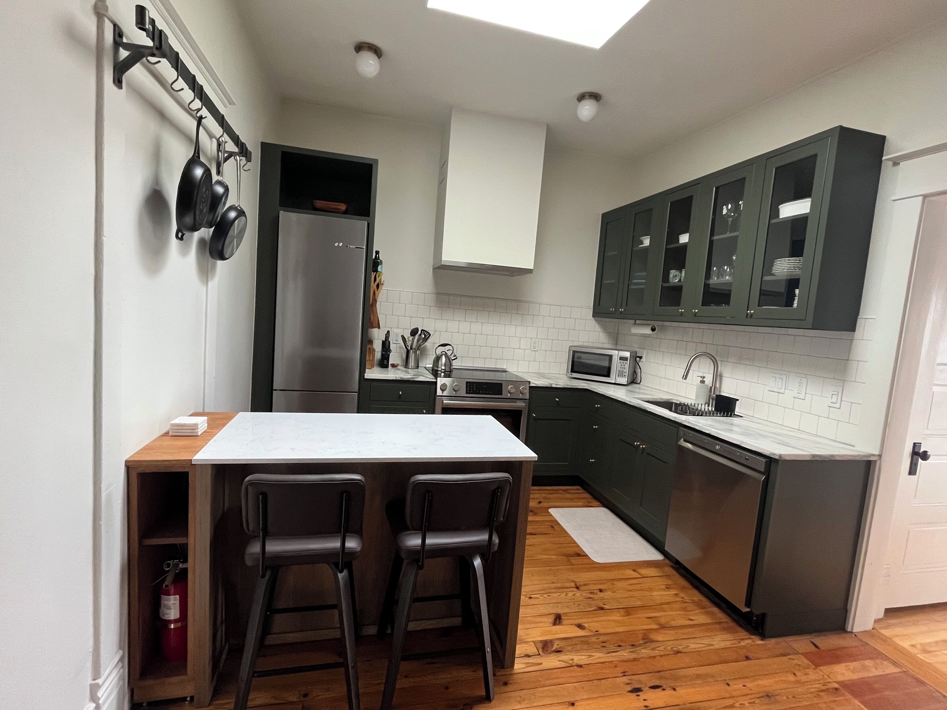 A modern kitchen with a central island, bar stools, stainless steel appliances, and dark green cabinets.