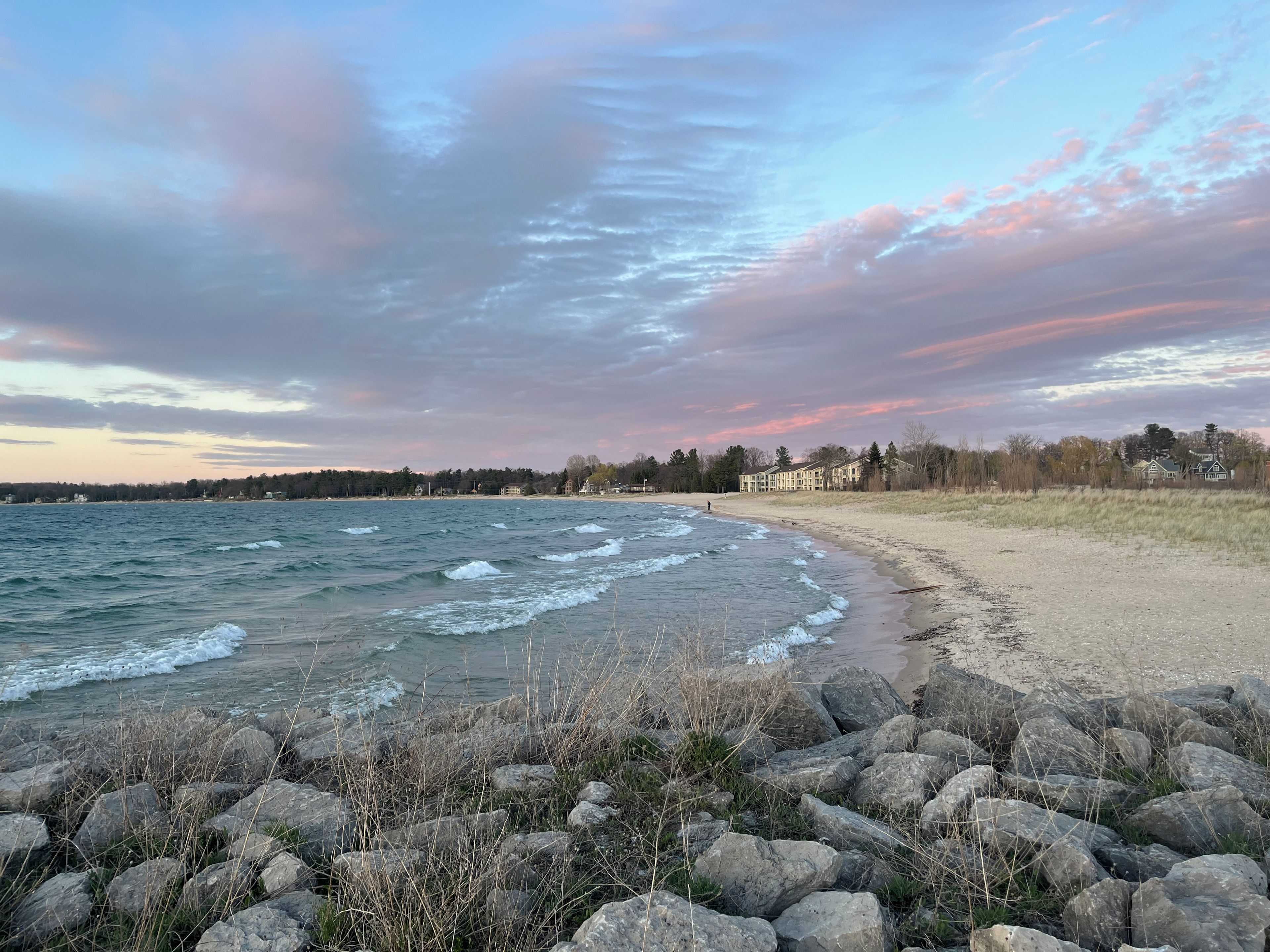 A beach with waves, a rocky foreground, and a row of buildings in the background.