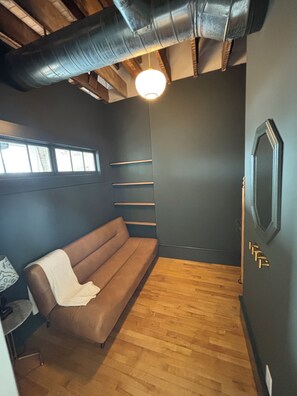 Ein Zimmer mit einem braunen Sofa, Holzboden, einer Wandregal und einer einzelnen Lampe.