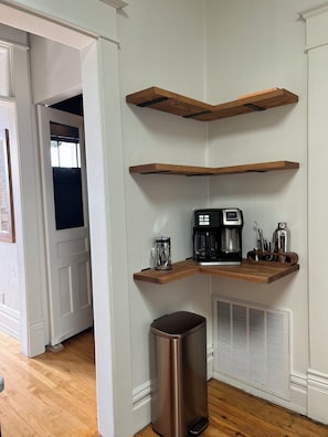 A corner of a room with a wooden shelf, a coffee maker, a thermos, and a trash can.