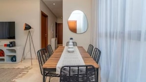 A dining area with a long wooden table, black chairs, a white table runner, a round mirror on the wall, and a floor lamp. - Prestigia Golf |Luxury 3BR| Pool&Atlas view (Marrakesh)