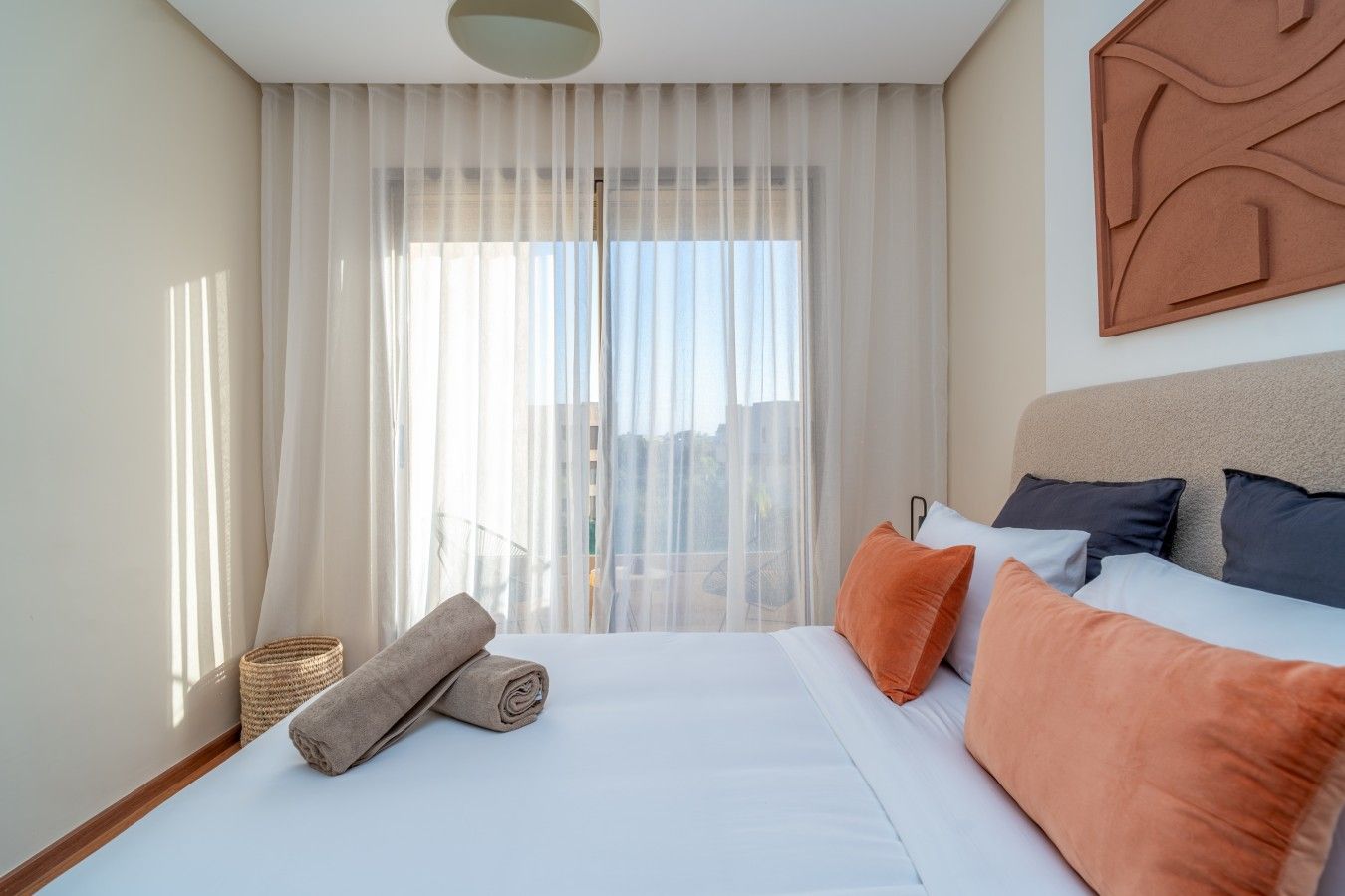 A hotel room with a bed, pillows, a rolled towel, a window with sheer curtains, and a city view.