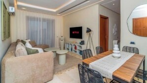 A modern living room with a sofa, coffee table, TV, and dining area. - Prestigia Golf |Luxury 3BR| Pool&Atlas view (Marrakesh)