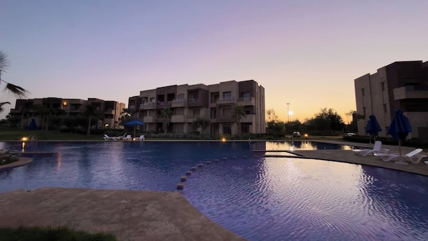 A swimming pool with lounge chairs and umbrellas, surrounded by multi-story buildings. - Prestigia Golf |Luxury 3BR| Pool&Atlas view (Marrakesh)