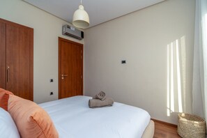 A bedroom with a bed, a wooden wardrobe, an air conditioning unit, a window with curtains, and a small woven basket. - Prestigia Golf |Luxury 3BR| Pool&Atlas view (Marrakesh)