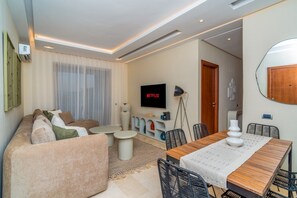 A modern living room with a sofa, coffee table, dining table, and chairs. A flat-screen TV is mounted on the wall. - Prestigia Golf |Luxury 3BR| Pool&Atlas view (Marrakesh)
