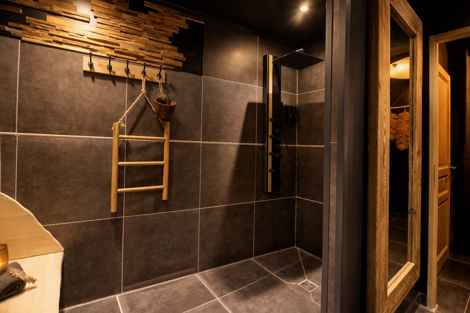 Un baño moderno con azulejos oscuros, una escalera de madera, una ducha con cabezal cuadrado y una puerta de cristal que da a un armario.