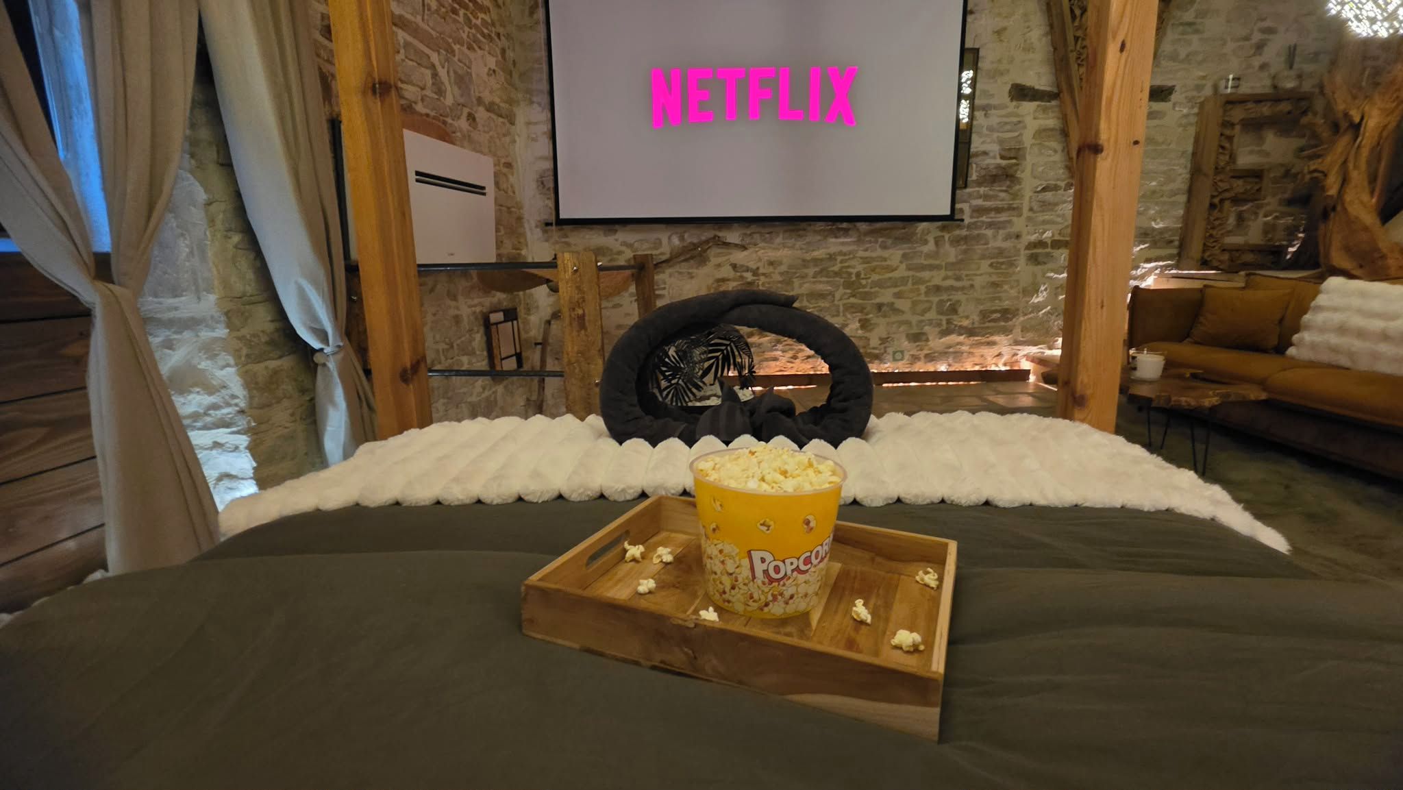 Ett mysigt rum med en säng, ett träbricka med popcorn och en skärm som visar ”NETFLIX”.