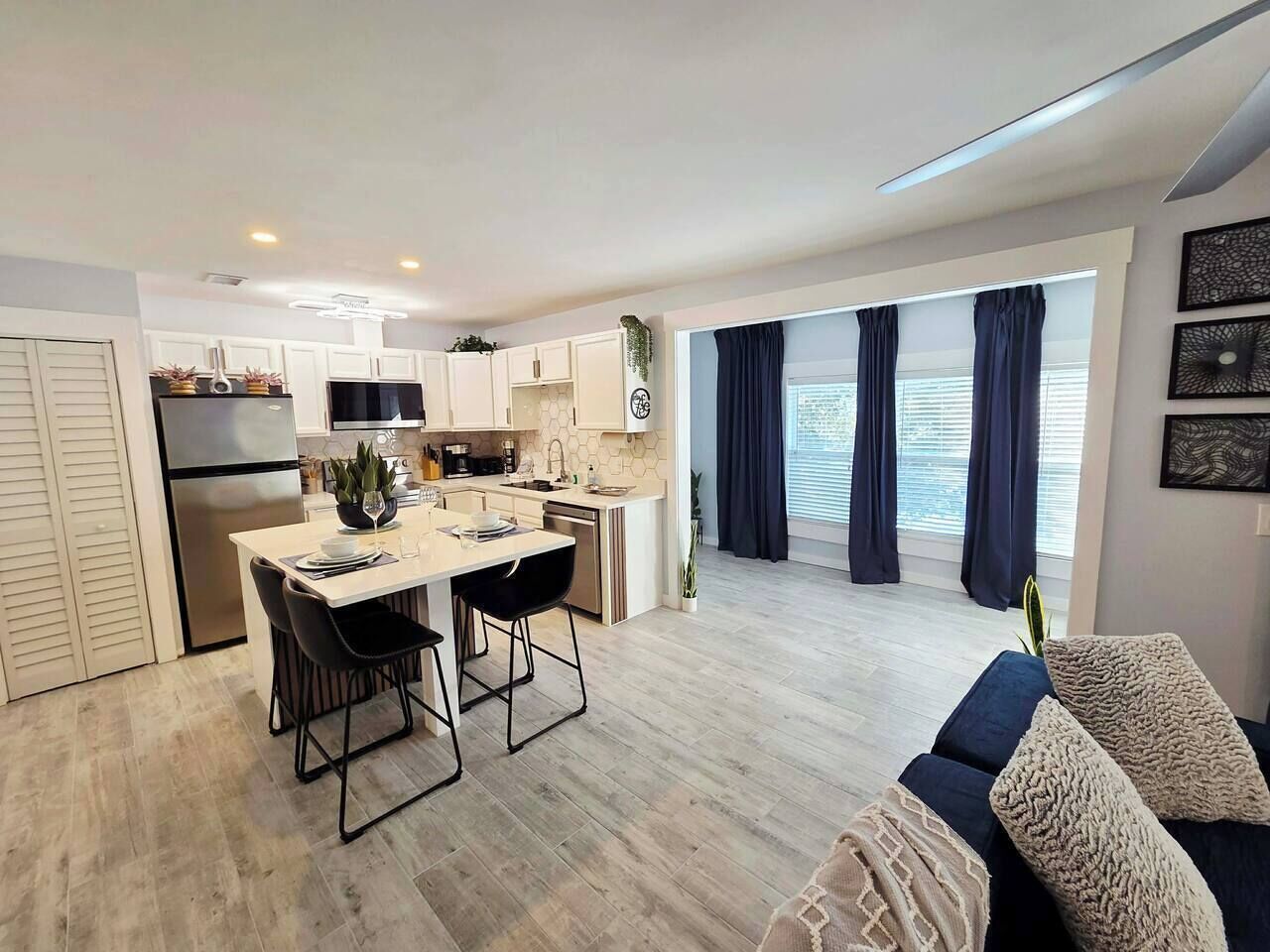 A modern kitchen with white cabinets, a dining area with a table and chairs, and a living area with a sofa and cushions.