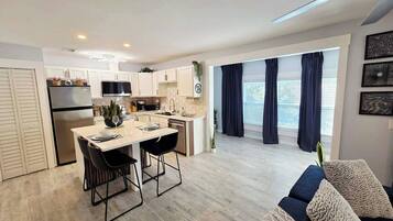 A modern kitchen with white cabinets, a dining area with a table and chairs, and a living area with a sofa and cushions.