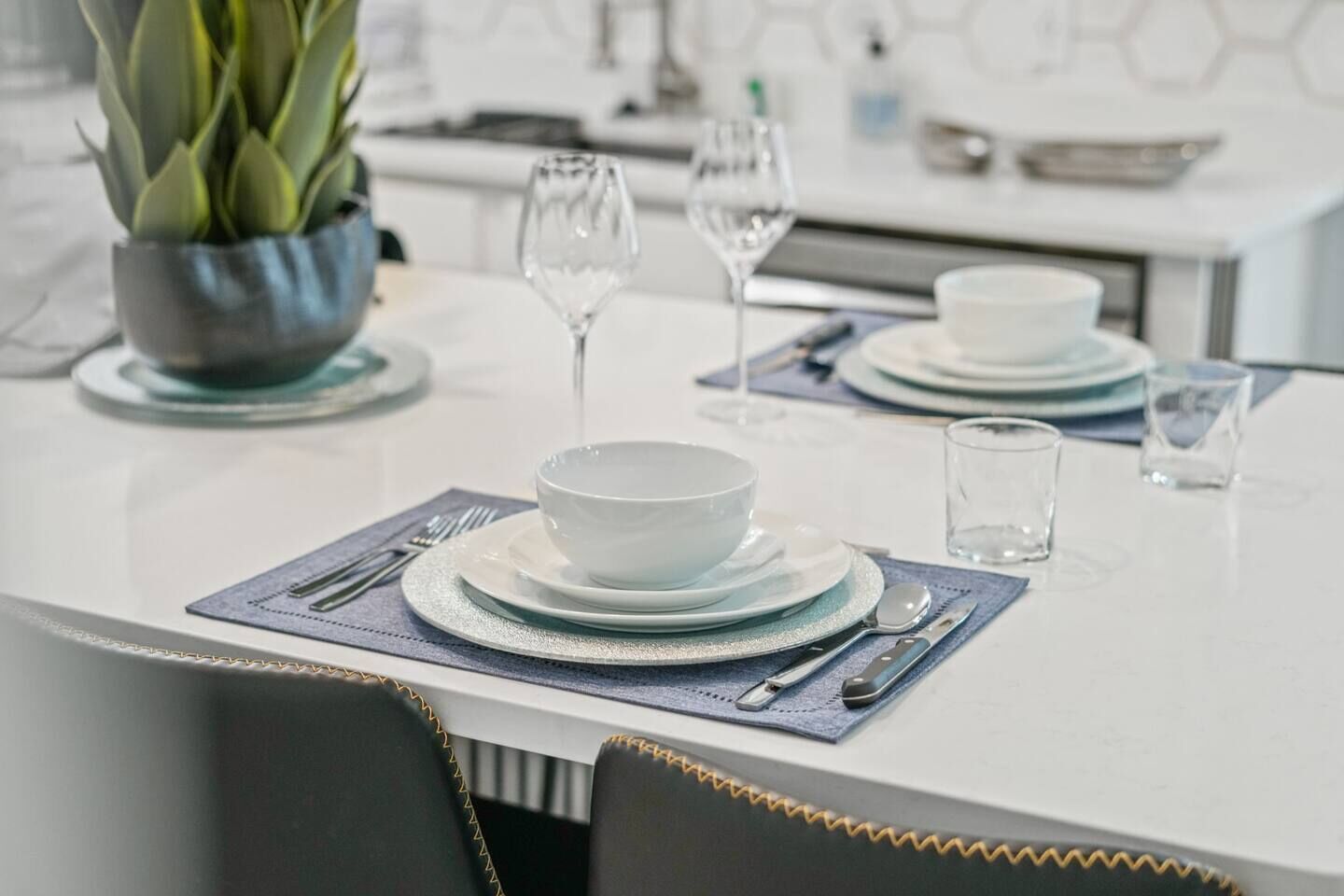 A modern dining area with a white table set for two, including plates, bowls, cutlery, and glasses.