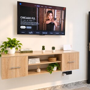 A flat-screen TV mounted on a wall displaying a show titled 'CHICAGO P.D.' with a subtitle about the show. Below the TV, there is a wooden console with books, a potted plant, and a small white device.