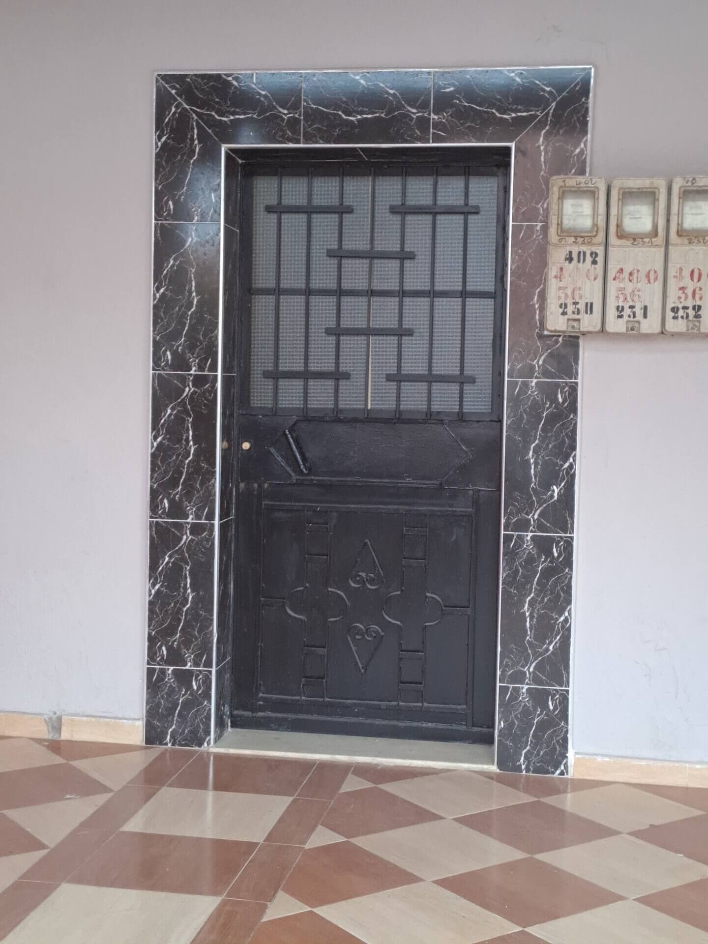 Puerta negra con un diseño de cuadrícula y una sección inferior con motivos de corazón y cruz, sobre un suelo de baldosas con patrón geométrico.