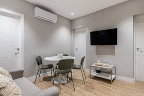Ein modernes Wohnzimmer mit rundem Tisch, grünen Stühlen, einem an der Wand montierten Flachbildfernseher und einem kleinen Beistelltisch mit einer Kerze.