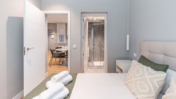 A modern bedroom with a bed, a bedside table, a nightstand, and a bathroom visible through an open door. - Waou Essence I Salamanca - Short-Term (Madrid)
