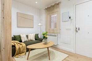 A room with a sofa, a coffee table, a radiator, a door, and a window with curtains. - Waou Cuatro Caminos II - Short-Term Rental (Madrid)