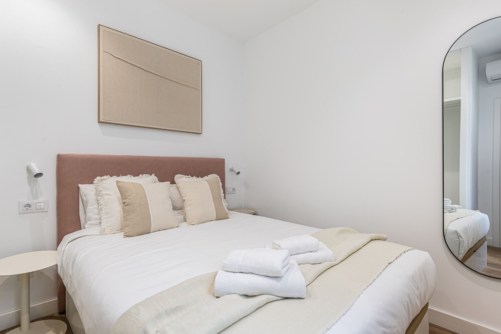 A neatly arranged hotel room with a bed, a bedside table, a wall-mounted light, a mirror, and a framed picture.