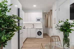 A compact kitchen with a built-in washing machine, a microwave, and a dining area with a glass table. - Waou El Rastro II - Short-Term Rental (Madrid)