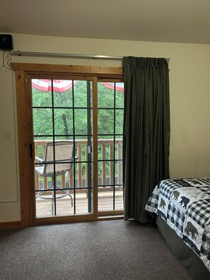 Ein Zimmer mit einer großen Schiebeglasfenster, das auf eine Terrasse im Freien führt, einem Bett mit kariertem Überwurf und einem Teppichboden.