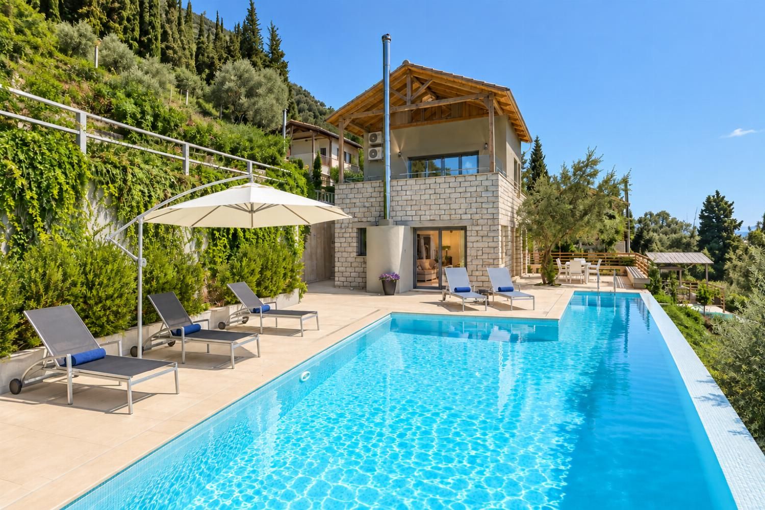 A modern villa with a swimming pool, lounge chairs, and a view of a forested hillside.