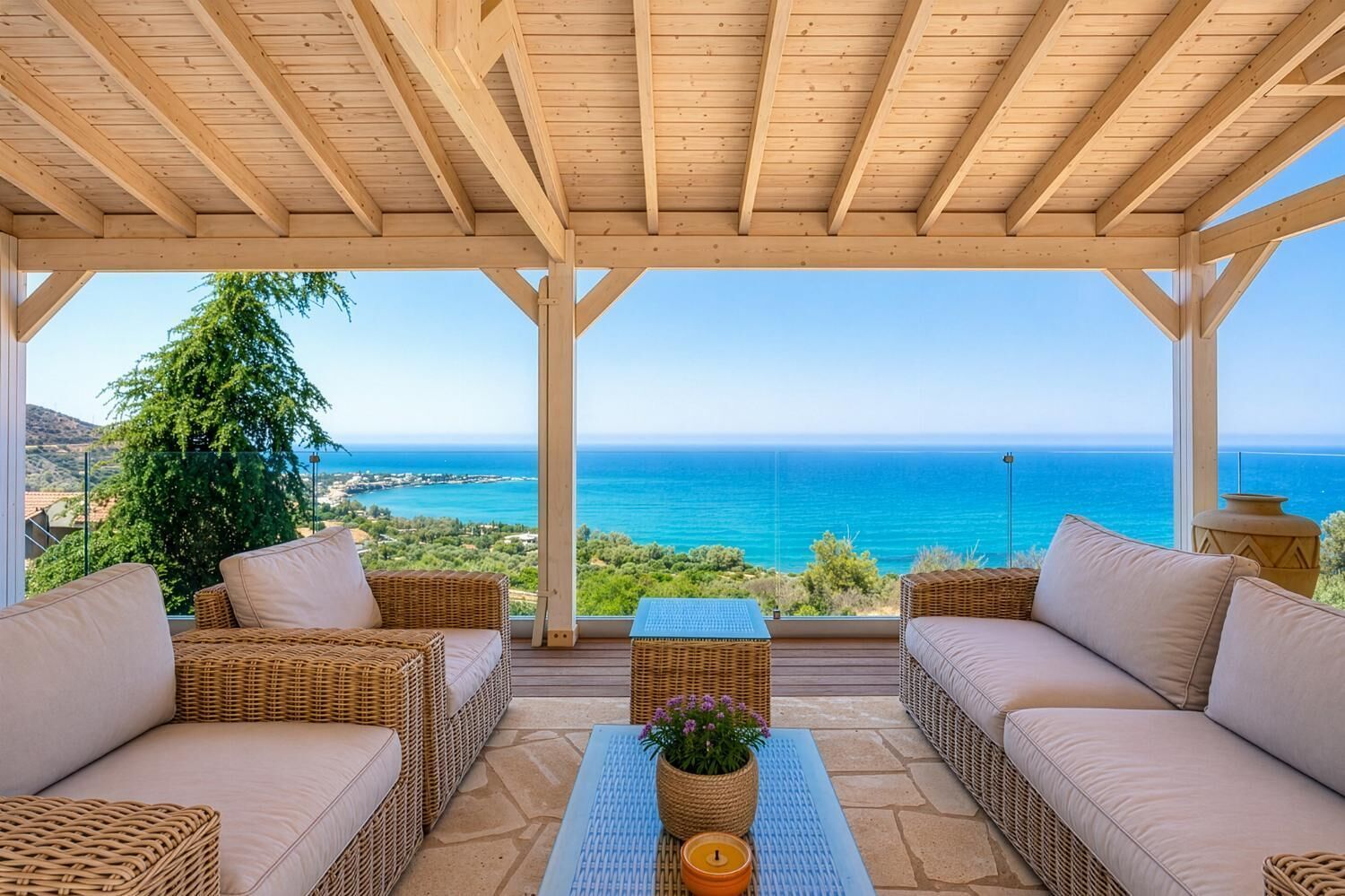 A covered outdoor seating area with wicker furniture, a central table with a plant, and a view of the sea and landscape.