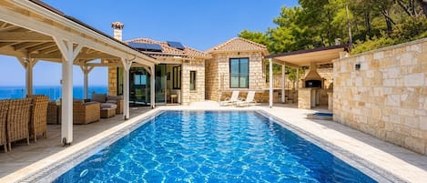 Une piscine agrémentée d’une pergola et de chaises longues, un bâtiment en pierre doté d’une cheminée, et une vue sur la mer.