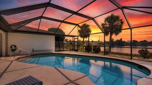 A swimming pool with a glass enclosure, surrounded by a view of a sunset over a body of water and palm trees. - Relaxing 3BR Lakehouse with Heated Pool & Lake Views (Port Charlotte)