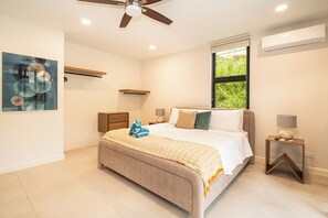 Ein Schlafzimmer mit einem Bett, Nachttischen, einem Deckenventilator, einem Fenster mit Aussicht und einer Wand-Klimaanlage.