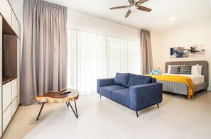 A modern hotel room with a sofa, a bed with a yellow blanket, a ceiling fan, and a painting on the wall. - Villa LaLa Land • 13 Suites • 2 Pools • Party Room (Jaco)