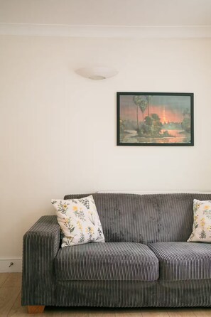 A room with a grey sofa, two pillows with floral patterns, and a framed painting of a landscape with a tower. - Cosy One-Double room flat near Big Ben (London)