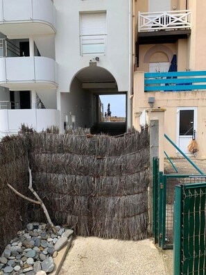 A gated area with a concrete pathway, a metal gate, and a building with a balcony and a window with blinds. - Jonquille Studio - Le Patio Residence (Mimizan)