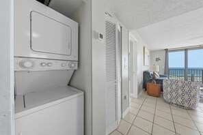 A laundry room with a front-loading washer and dryer, a door leading to a balcony with a view of the beach, and a patterned ottoman on the balcony. - 🏖️ Seaside Beach + Racquet 3801 | Gulf Front Corner Condo + Wrap Balcony (Orange Beach)