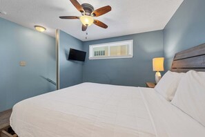 Ein Schlafzimmer mit einem Bett, einem Deckenventilator, einem Fernseher, einem Fenster mit Jalousien und einem Nachttisch mit Lampe.