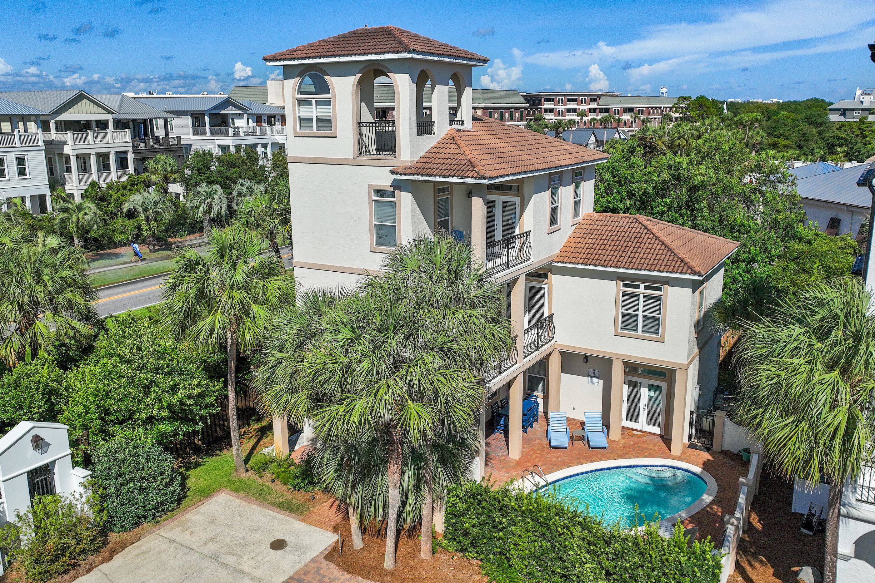 A multi-story residential building with a swimming pool, surrounded by lush greenery and other houses.