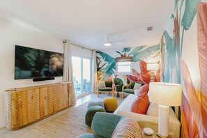 A modern living room with a flat-screen TV, a wooden cabinet, colorful cushions, and a large tropical-themed wall mural. - Welcome to The Sunnyside Pool House! Your Ultimate Coastal Escape (Sunnyside)