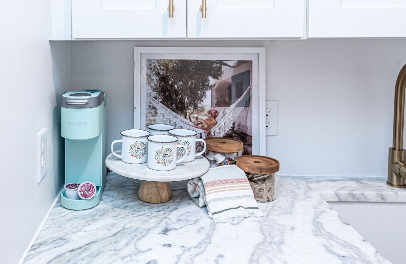 A kitchen with a coffee maker, a tea kettle, a tray with mugs, a small wooden table, and a newspaper dispenser.