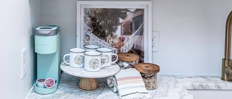 A kitchen with a coffee maker, a tea kettle, a tray with mugs, a small wooden table, and a newspaper dispenser.