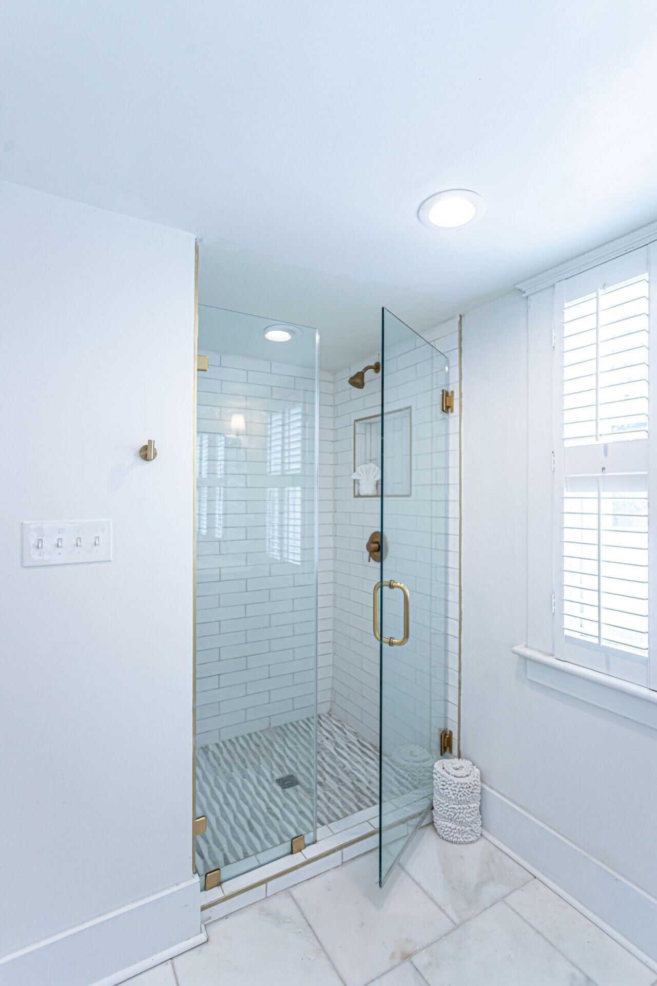 A modern bathroom with a glass-enclosed shower, white tiled walls, and a window with blinds.