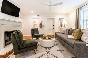 Una sala de estar con chimenea, un sofá, un sillón, una mesita de centro y un televisor montado en la pared.