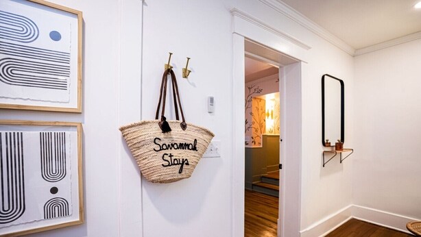 Ein Flur mit einem gerahmten abstrakten Kunstwerk, einer gewebten Tasche mit dem Schriftzug 'Savannah Stays' und einem Spiegel an der Wand.