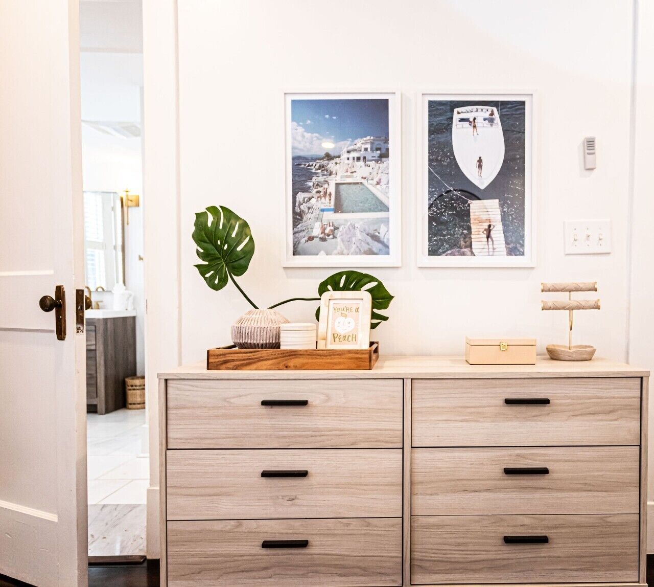 A hotel room with a wooden dresser, two framed pictures on the wall, a plant, and a small wooden box on the dresser.