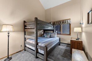 Un lit superposé avec une couvre-lit à motifs, une lampe de chevet, une petite table de nuit en bois et une fenêtre avec des rideaux.