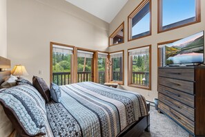 Ein Schlafzimmer mit Bett, Schminktisch, Fernseher und großen Fenstern mit Blick auf Grünflächen.