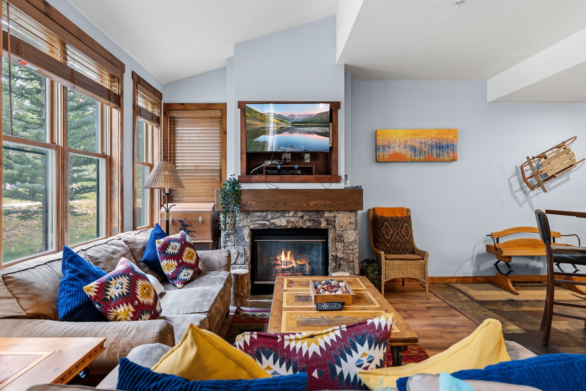 A cozy living room with a fireplace, a television mounted above it, and a large window with a view of trees.