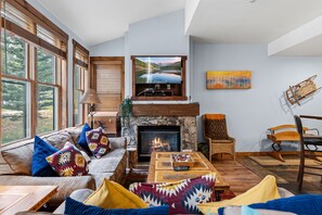 A cozy living room with a fireplace, a television mounted above it, and a large window with a view of trees. - Ideally Located to Downtown 1 Bedroom, 1 Bath in Main Street Junction (Breckenridge)