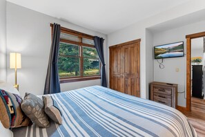 Un dormitorio con una cama, una ventana con cortinas, un armario de madera, un televisor en la pared y un mueble pequeño.